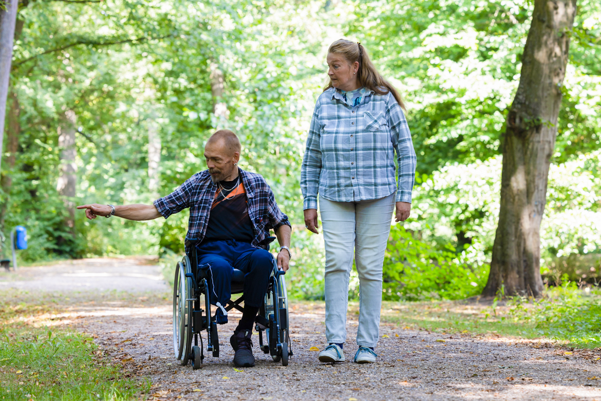 Zwei Personen gehen im Park spazieren. Der Mann im Rollstuhl zeigt auf etwas. Die Dame neben ihm schaut interessiert in die gezeigte Richtung.