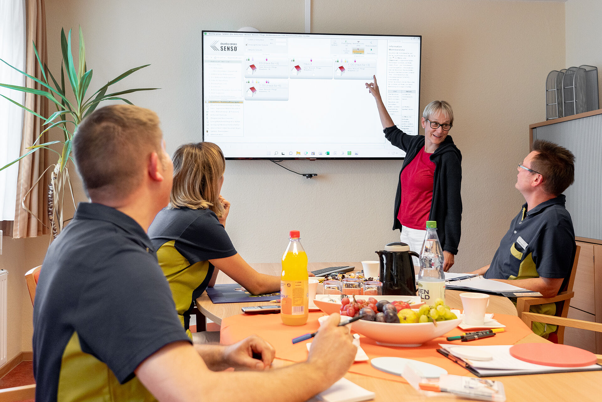 Vier Pflegekräfte sitzen an einem Tisch in einem Meeting. Im Hintergrund steht eine Frau an einem Bildschirm und präsentiert. 