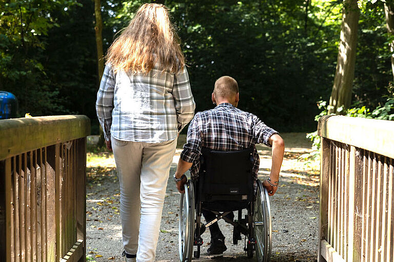 Eine Frau geht über eine Brücke, in einem Park. Neben ihr fährt ein Mann im Rollstuhl. Beide sind nur von hinten zu sehen.