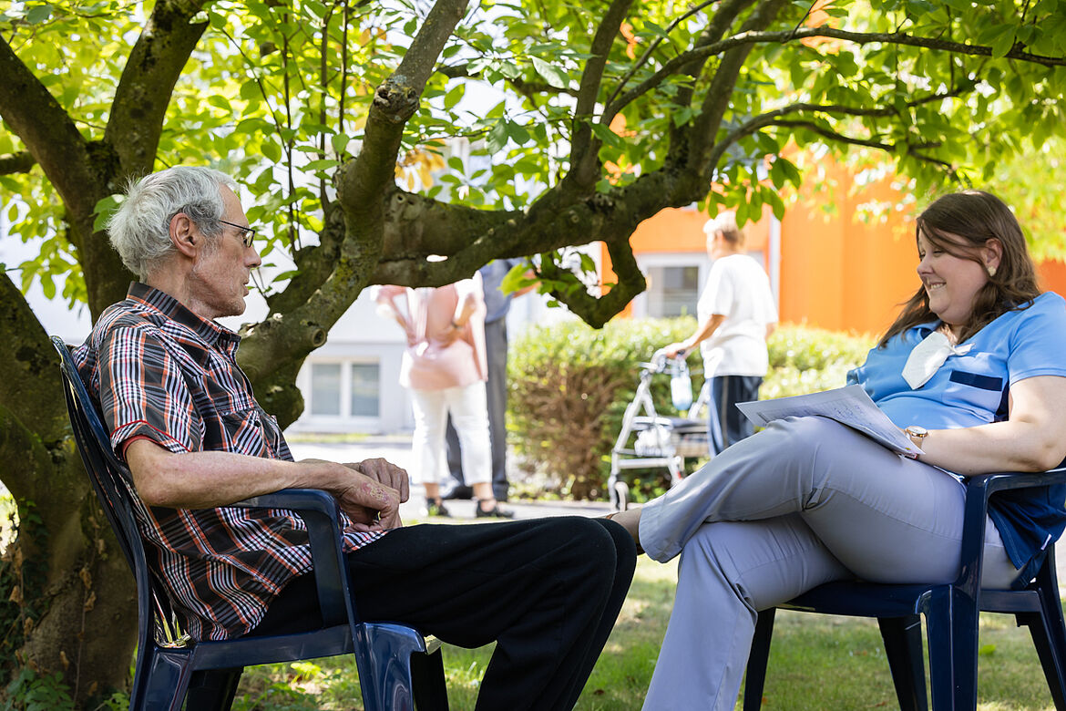 Ein älterer Mann und eine junge Pflegekraft sitzen sich unter einem Baum gegenüber.