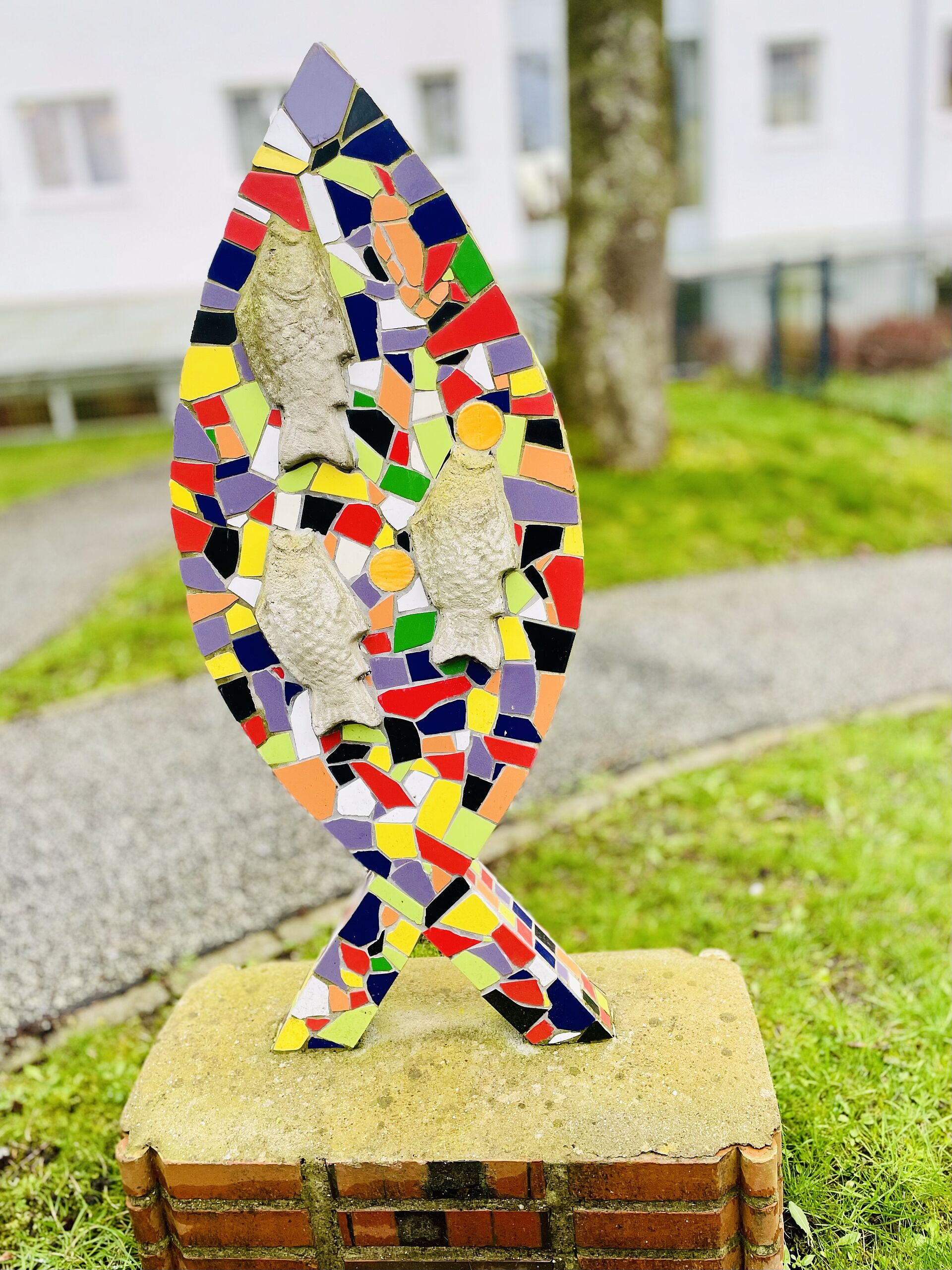 Ein Fisch aus Mosaiksteinen steht unter einem Baum, auf dem Gelände des Reha-Hauses.